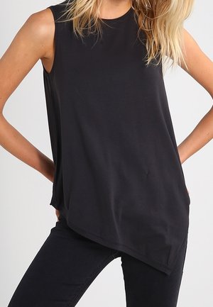 Femme portant un top noir asymétrique sans manches avec un côté plus long, associé à un pantalon noir, mains sur les hanches.