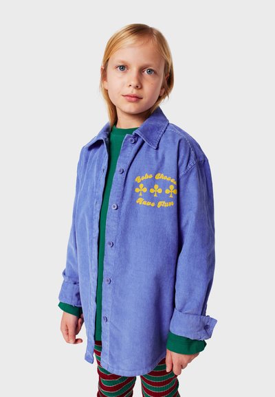 Camicia di velluto a coste viola chiaro con colletto, chiusura a bottoni e ricamo di fiori gialli. Indossata sopra una maglietta verde e leggings a righe.