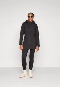 Schwarze Kapuzenjacke mit Reißverschluss und zwei Fronttaschen, kombiniert mit schwarzen Leggings und hellen Sneakers. Modell trägt eine Sonnenbrille.