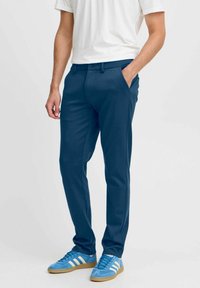 Moški nosi temno modre oprijete hlače s stranskimi žepi in svetlo modre superge Adidas z belimi črtami, stoji ob enem tonirnem ozadju.