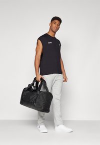 Bolsa de deporte negra con asas dobles. El modelo lleva una camiseta negra sin mangas y pantalones de chándal grises con zapatillas blancas. Diseño simple.