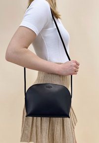 Black leather crossbody bag with a curved top, slim adjustable strap, and embossed logo on the front, showcased against a neutral background.