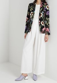 Blazer met bloemenpatroon in zwart en levendige kleuren, gecombineerd met witte wijde broek en lavendelkleurige block-heeled muilen.