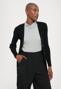 Black ribbed cardigan with gold buttons, worn over a light blue top. Styled with tailored black trousers, featuring a relaxed fit and side pockets.