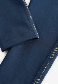 Tamno plave tajice izrađene od mekanog materijala. Imaju oznaku brenda "BAKER by TED BAKER" u zlatnoj boji sa strane. Glatka tekstura, prilagođeni dizajn.