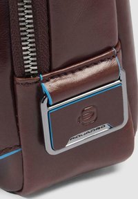 Borsa in pelle marrone con una cerniera argentata e un'etichetta logo lucida con accenti blu. Caratterizzata da una cucitura dettagliata e una superficie liscia.