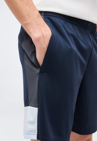 Άνδρας που φοράει ναυτικό μπλε αθλητικό σορτς με μαύρες και ανοιχτό γκρι πλευρικές λωρίδες, το χέρι του ακουμπάει στην δεξιά τσέπη, με φόντο ένα μονόχρωμο υπόβαθρο.