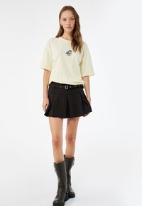 Valkoinen oversized t-paita, jossa on kirjailtu kuvio, yhdistetty mustaan pleated-minihameeseen, jossa on vyö. Korkeat mustat nahkasaappaat.