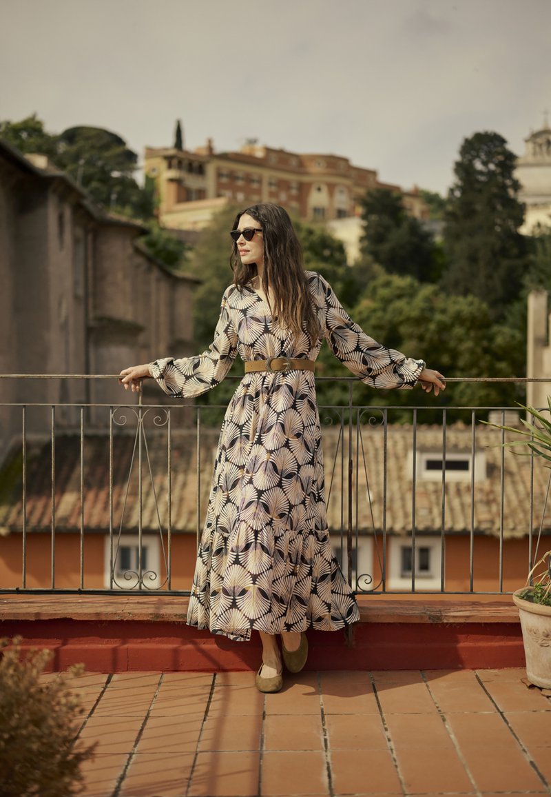 Langes Blumenkleid mit einem schwarzen und cremfarbenen Palmblattmuster, langen Ärmeln, tailliert mit einem braunen Gürtel, kombiniert mit olivgrünen Schuhen.