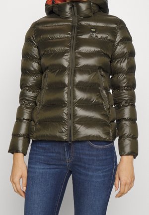 Kobieta ubrana w błyszczącą, oliwkowozieloną puchową kurtkę z kapturem, z przodu zapinaną na zamek, w połączeniu z niebieskimi dżinsami.