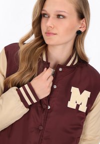 Bordowa i beżowa kurtka varsity z paskowanymi mankietami, zatrzaskami i haftowanym kremowym logo "M" na lewej piersi.