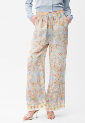 Frau trägt hellblaue, weit geschnittene Hosen mit aufwändigen orange-goldenen Blumenmustern, kombiniert mit goldenen offenen Sandalen mit Absatz und einem kurzen Cardigan.