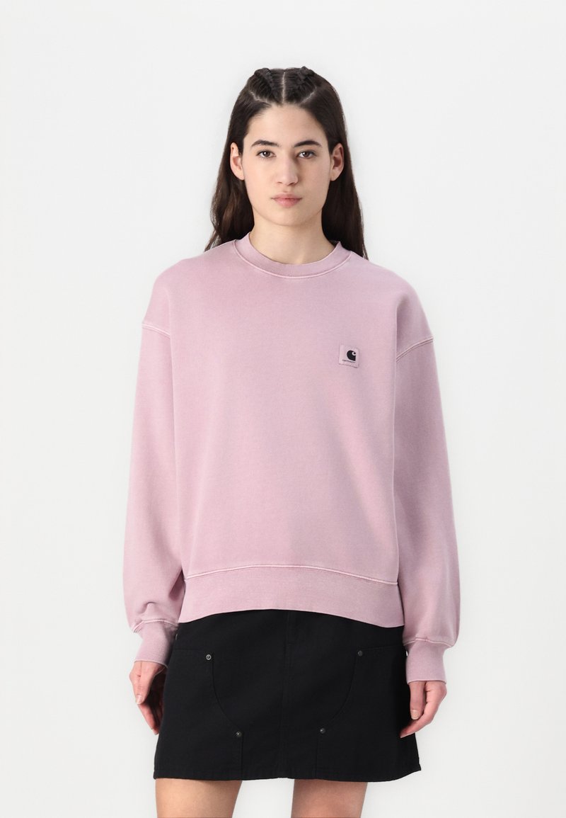 Ljusrosa sweatshirt med långa ärmar, ribbade muddarna och rund halsringning. Har en liten svart logotyppatch. Matchas med en svart kjol.