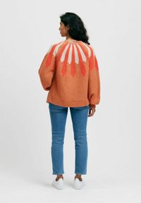 Oranssissa neulepaidassa on geometrijainen kuvio valkoisena ja korallina selässä. Paidassa on puffihihat ja rento malli.