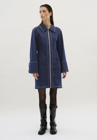 Jeansklänning i mörkblått med spetsig krage, dragkedja fram, långa ärmar och kontrasterande vita sömmar. Buren med svarta knähöga stövlar.