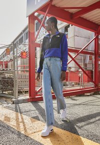 Giacca in pile nera e blu con zip, jeans a gamba larga blu chiaro e sneakers bianche con platfrom. Lo sfondo presenta elementi di costruzione.