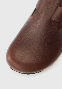 Bruine leren schoen met een gestructureerd oppervlak, ronde neus en uitsnijdingsdetails. Zwarte buitenzool met een eenvoudig ontwerp voor extra grip.