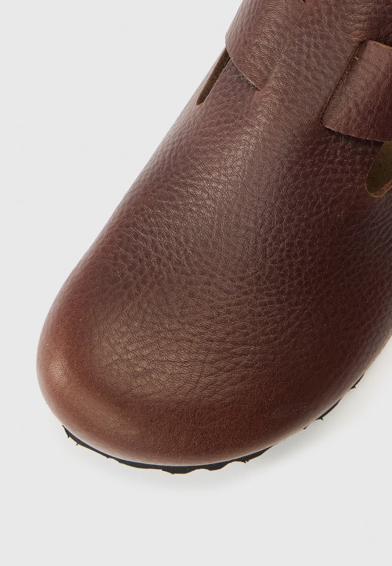 Scarpa in pelle marrone con superficie texturizzata, punta arrotondata e dettagli ritagliati. Suola nera con design semplice per una maggiore aderenza.