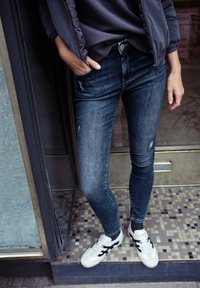 Kapeavat farkut tummansinisestä denimmateriaalista, jossa on lievää kulutusta, yhdistettyinä valkoisiin tennareihin, joissa on mustia yksityiskohtia. Päällä harmaa neule ja takki.
