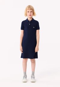 Mekko navy sinisellä värillä, jossa on lyhyet hihat ja klassinen kaulus. Mekossa on pieni vasemmalla rintapuolella sijaitseva tasku ja vihreä logotehoste.