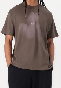 Hnědé bavlněné tričko s fialovým designem připomínajícím oblak, kulatým výstřihem a malým bílým logem Nike na hrudi. Volný střih a krátké rukávy.