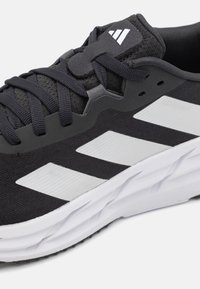 Chaussure de sport noire avec une tige texturée, un logo à trois bandes blanches et une semelle blanche rembourrée avec un motif ondulé.
