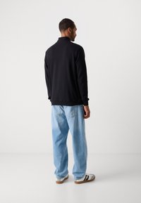 Hombre de espaldas vistiendo una camiseta negra de manga larga, jeans azul claro y zapatillas blancas frente a un fondo liso de color gris claro.