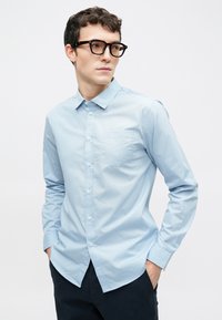 Chemise bleu clair à boutons avec un motif discret de points, manches longues et une poche poitrine unique. Texture lisse et col structuré.