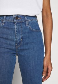 Jeans de mezclilla azul de tiro alto con una forma ajustada, con costuras doradas, cierre de botón en la parte frontal y una superficie lisa, ligeramente texturizada.