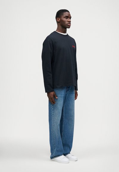 Egy férfi áll, fekete, hosszú ujjú pólót viselve, piros felirattal, laza kék farmernadrágban és fehér sportcipőben, egy egyszínű világos háttér előtt.