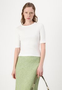 Biely rebrovaný top s krátkym rukávom a okrúhlym výstrihom v kombinácii s textúrovanou zelenou sukňou. Modelka drží zelenú kabelku s popruhom.