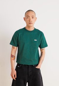Tricou din bumbac verde, cu decolteu rotund și mâneci scurte. Are un mic logo alb pe piept. Asortat cu pantaloni din denim negri.