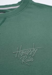 Grön bomullströja med ribbad rund halsringning och strukturerad broderad logotyp "Honesty Rules" i en något ljusare grön nyans.