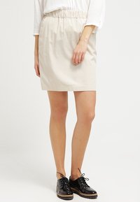 Jupe en coton beige avec une taille élastique, longueur mi-cuisse, texture lisse, accompagnée de chaussures noires à lacets.