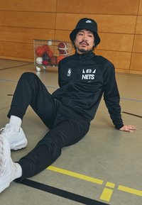 Tuta nero con logo NBA, dettagli bianchi e cappello a secchiello. Include scarpe da ginnastica e si trova su un pavimento da palestra con linee colorate.