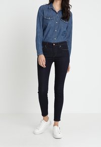 Camisa de mezclilla con botones y dos bolsillos en el pecho, combinada con jeans ajustados oscuros y zapatillas blancas, sobre un fondo liso.