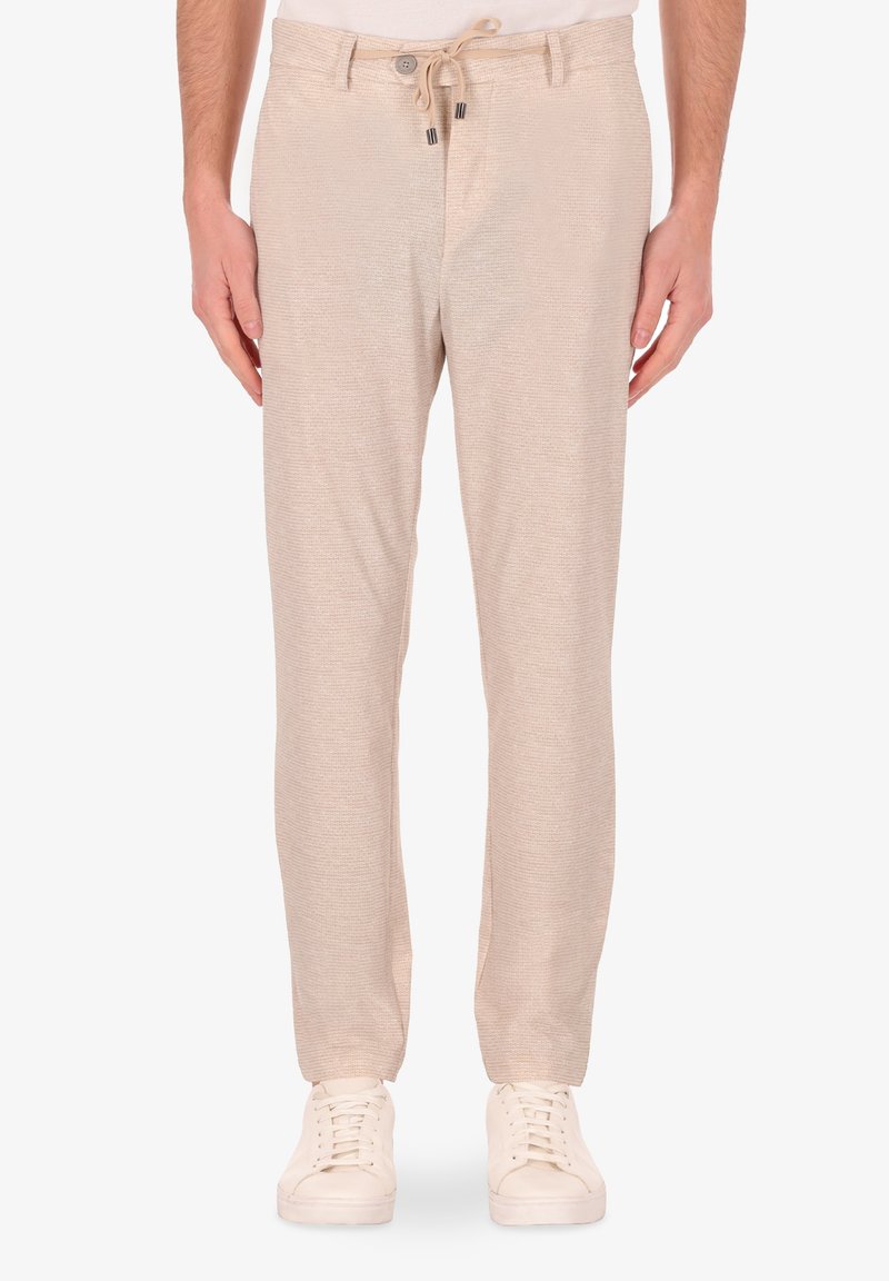 Pantaloni intrecciati beige con vita elastica e lacci, dotati di due tasche laterali e un taglio slim. Abbinati a sneaker bianche.