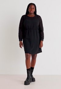 Robe noire avec des manches longues en dentelle, taille cintrée et une légère forme en A. Portée avec des bottes à plateforme noires. Tissu uni, design décontracté.