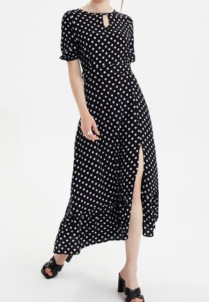 Schwarzes Midikleid mit weißen Polka-Dots, kurzen Ärmeln, Ausschnitt mit Schlüsselloch, Oberschenkelschlitz, kombiniert mit schwarzen Sandalen mit Absatz und silberner Kettenkette.