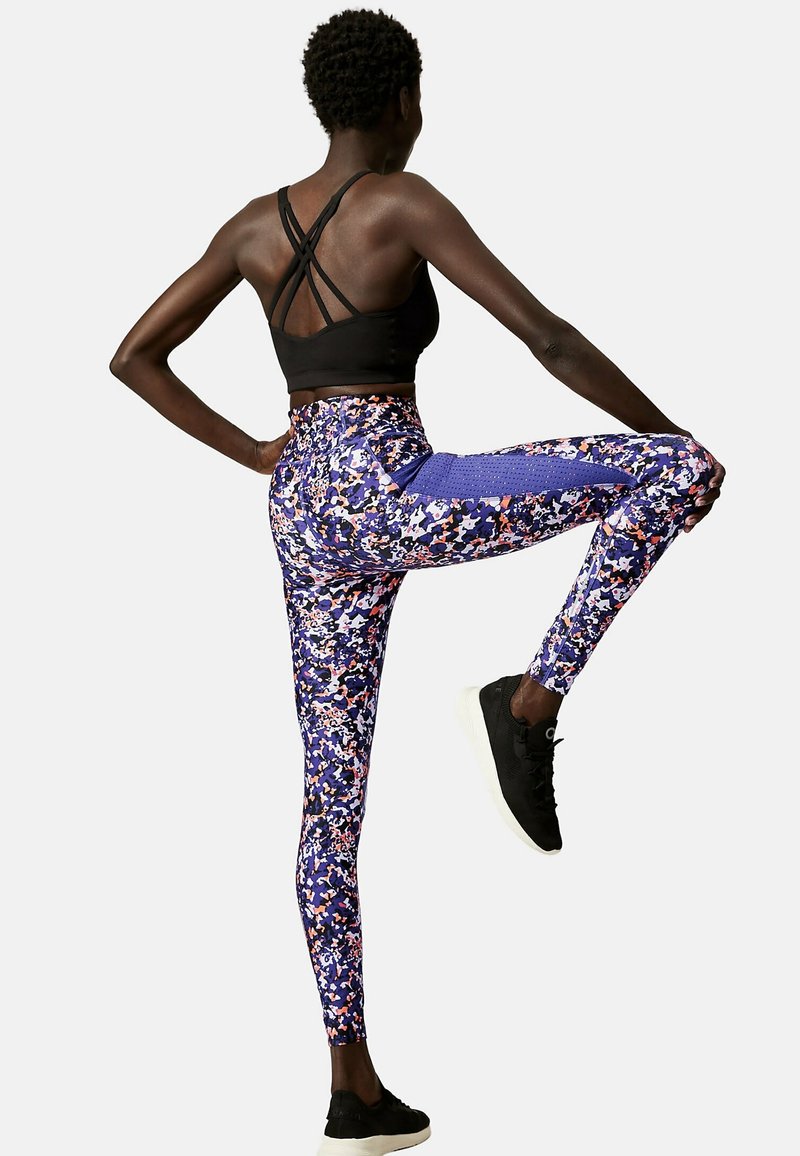 Soutien-gorge de sport noir avec des bretelles croisées, associé à un legging vibrant à motifs comportant des nuances de violet, rose et blanc, avec un tissu texturé.