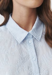 Blusa azul claro con patrones florales bordados, un cuello clásico y botones. La tela parece suave y con textura.