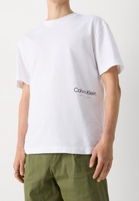Biała bawełniana koszulka z okrągłym dekoltem i krótkimi rękawami, z czarnym logo "Calvin Klein" wydrukowanym po lewej stronie u dołu.