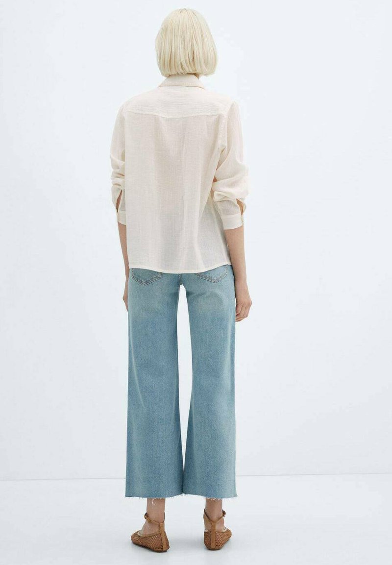 Camicia beige chiaro, morbida e con maniche arrotolate, abbinata a jeans in denim azzurro chiaro a gamba larga. Lo sfondo è neutro.
