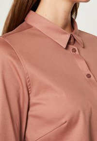 Femme portant une chemise en satin rose poussiéreux à manches longues, avec un col pointu et trois boutons visibles sur la patte de boutonnage avant.