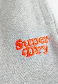 Šedá tkanina s jemnou textúrou, zdobená výrazným oranžovým vyšitým textom "Super Dry". Žiadne ďalšie detaily nie sú viditeľné.