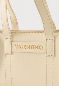 Bež usnjena torbica z zgornjimi ročaji, opremljena z zlatim logotipom in zadrgo. Teksturirana površina z vidnimi šivi.