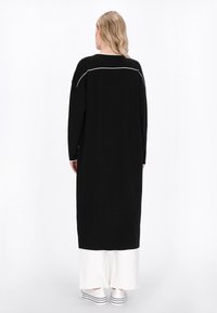 Schwarzes langärmeliges Kleid mit einer weißen Akzentlinie hinten. Lockerer Schnitt und leicht strukturierter Stoff, kombiniert mit weißen, schichtweise gestalteten Hosen und Sneakers.