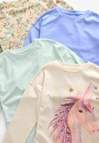 Trois tops à manches longues : un à motifs floraux, un bleu clair, un vert menthe. Le haut crème présente un design de licorne avec des sequins.