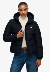 Mørkeblå pufferjakke med lynlås og hætte, der har elastiske manchetter og et lille hvidt logo på brystet. Båret over en beige top.