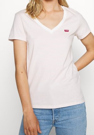 Kobieta nosząca białą i bladoróżową koszulkę w paski z krótkim rękawem i dekoltem w serek z czerwonym logo Levi's oraz niebieskie dżinsy z ręką w kieszeni.
